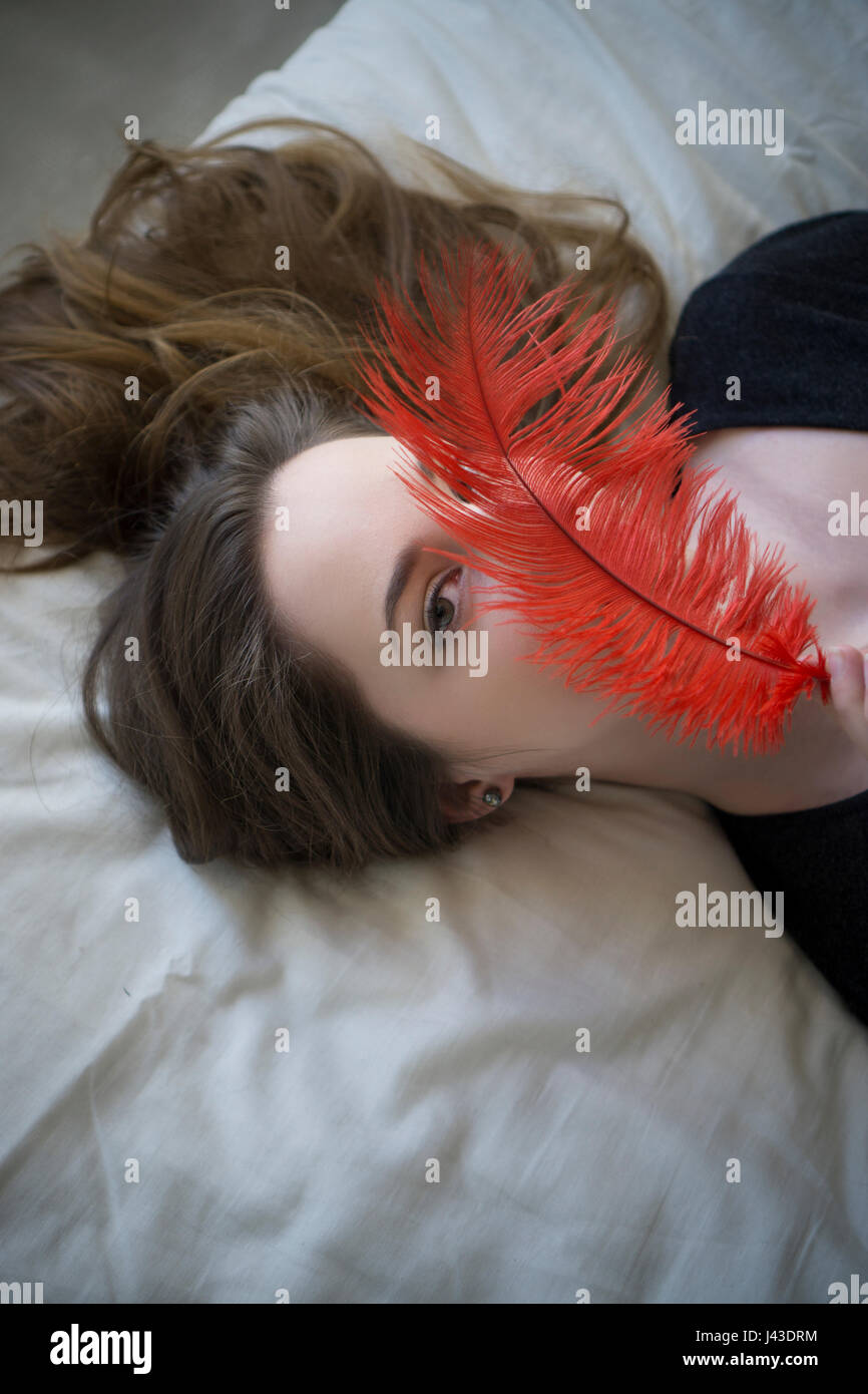 Beautiful young woman laying down in bed hiding face with a red feather
