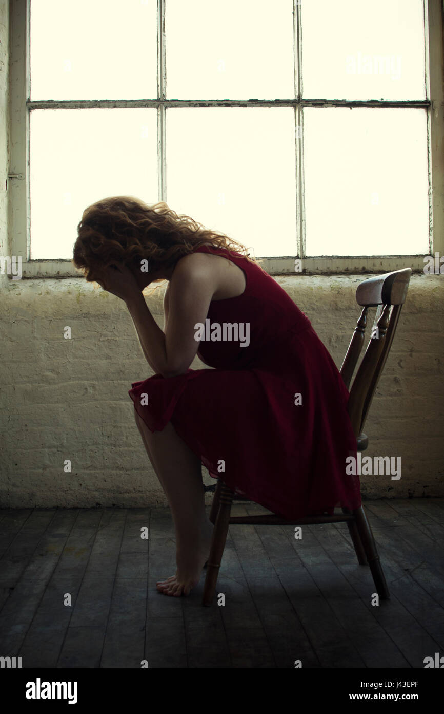 Redhead woman sitting on a chair by the window hiding face with hands crying Stock Photo