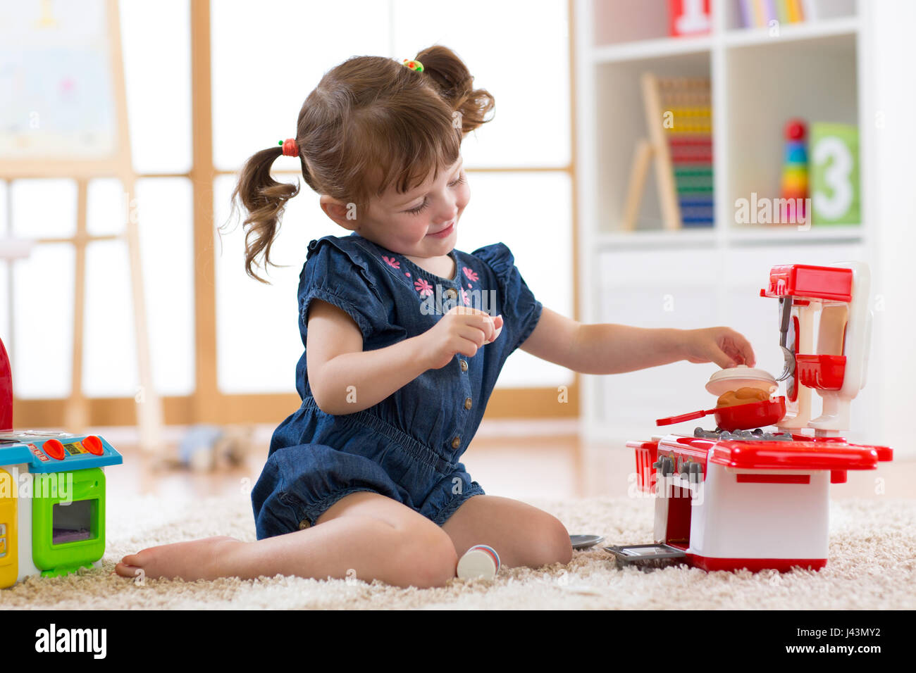 Little cute girl playing with utensil toys. Toddler kid in a playroom. Kid sitting on floor and cook in toy kitchen. Stock Photo