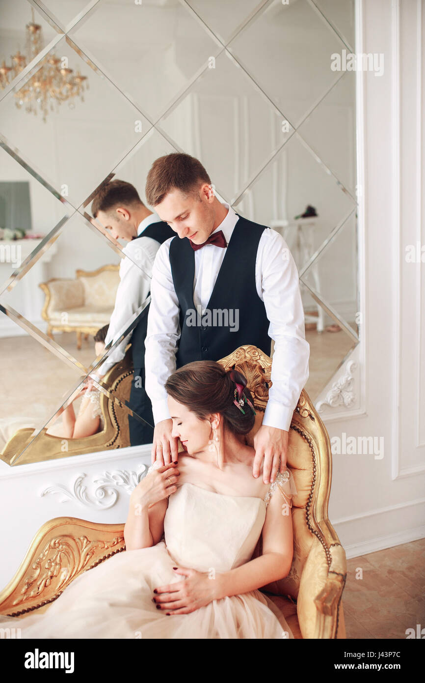 Bride in beautiful dress and groom in black suit sitting on sofa ...