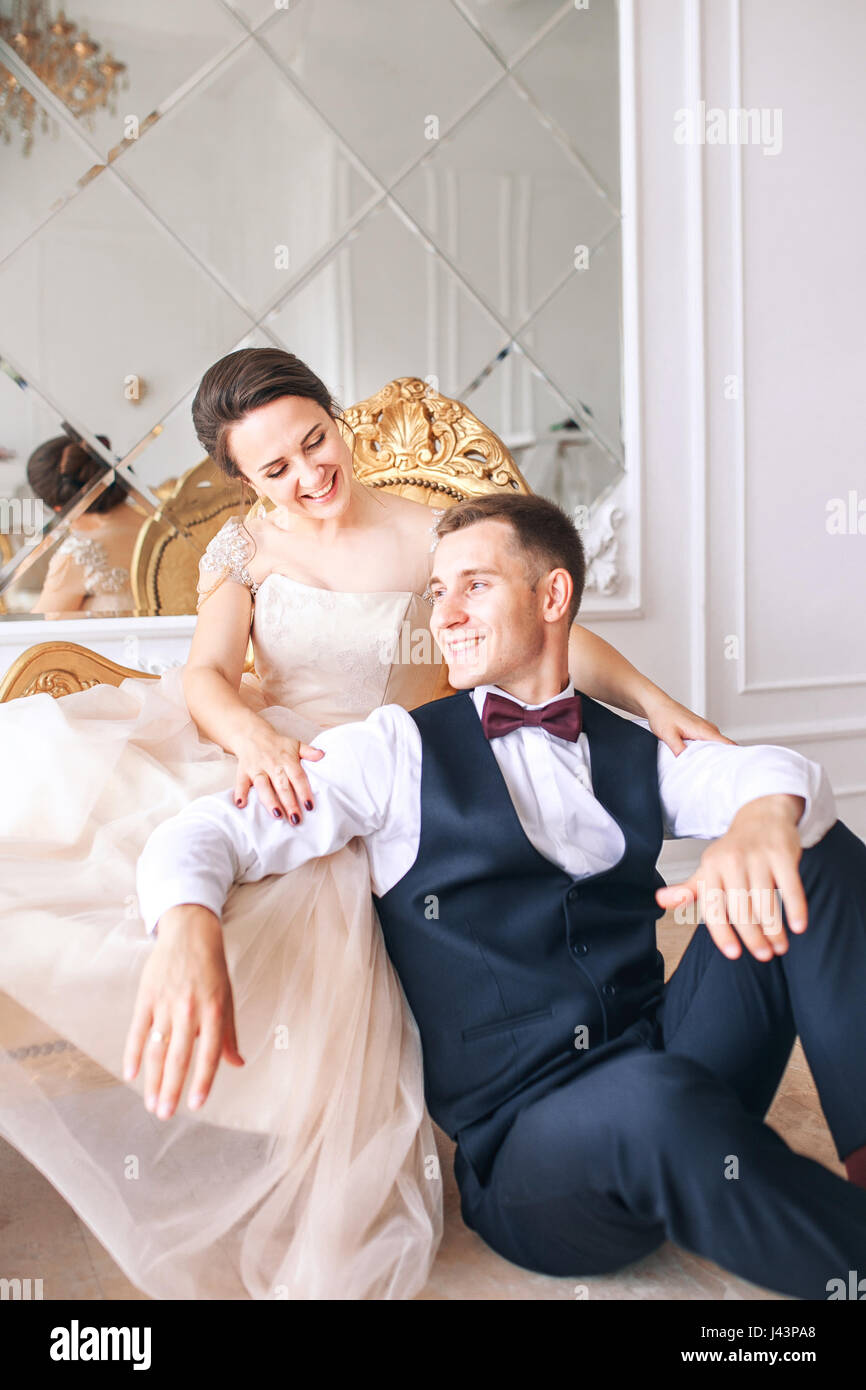 Bride in beautiful dress and groom in black suit sitting on sofa ...
