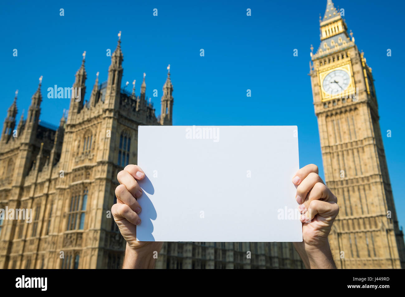 Hands holding blank sign in front of the Houses of Parliament at Westminster Palace with Big Ben in bright blue sky in London, England Stock Photo