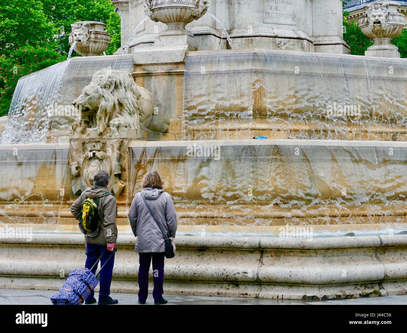 Couple wearing rain jackets and pulling rolling suitcase looking at Fountain Saint-Sulpice in light rain. Paris, France Stock Photo