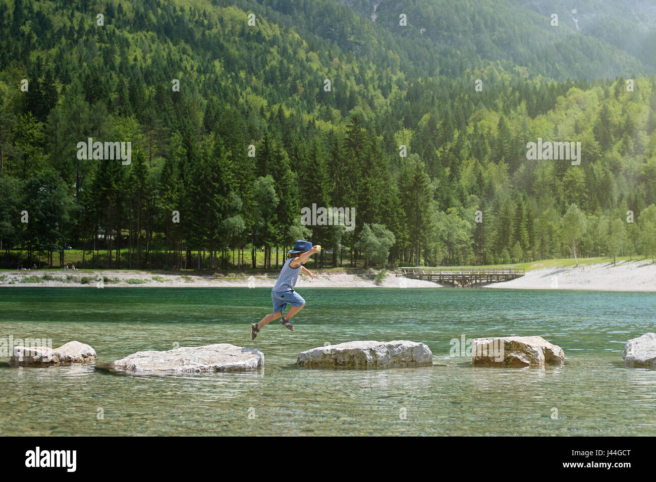 Adorable young boy with a hat crossing river or water jumping from rock to rock. Crossing the gap, freedom, liberation, success, avoiding danger, cour Stock Photo