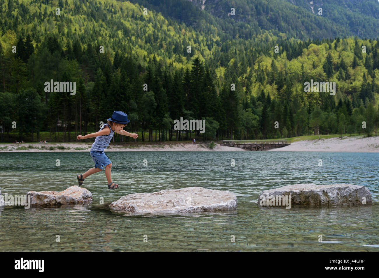 Adorable young boy with a hat crossing river or water jumping from rock to rock. Crossing the gap, freedom, liberation, success, avoiding danger, cour Stock Photo