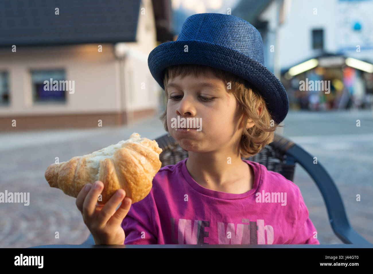 Cute little boy with blue hat sitting at the table looking at tasty croissant he is eating. Stock Photo