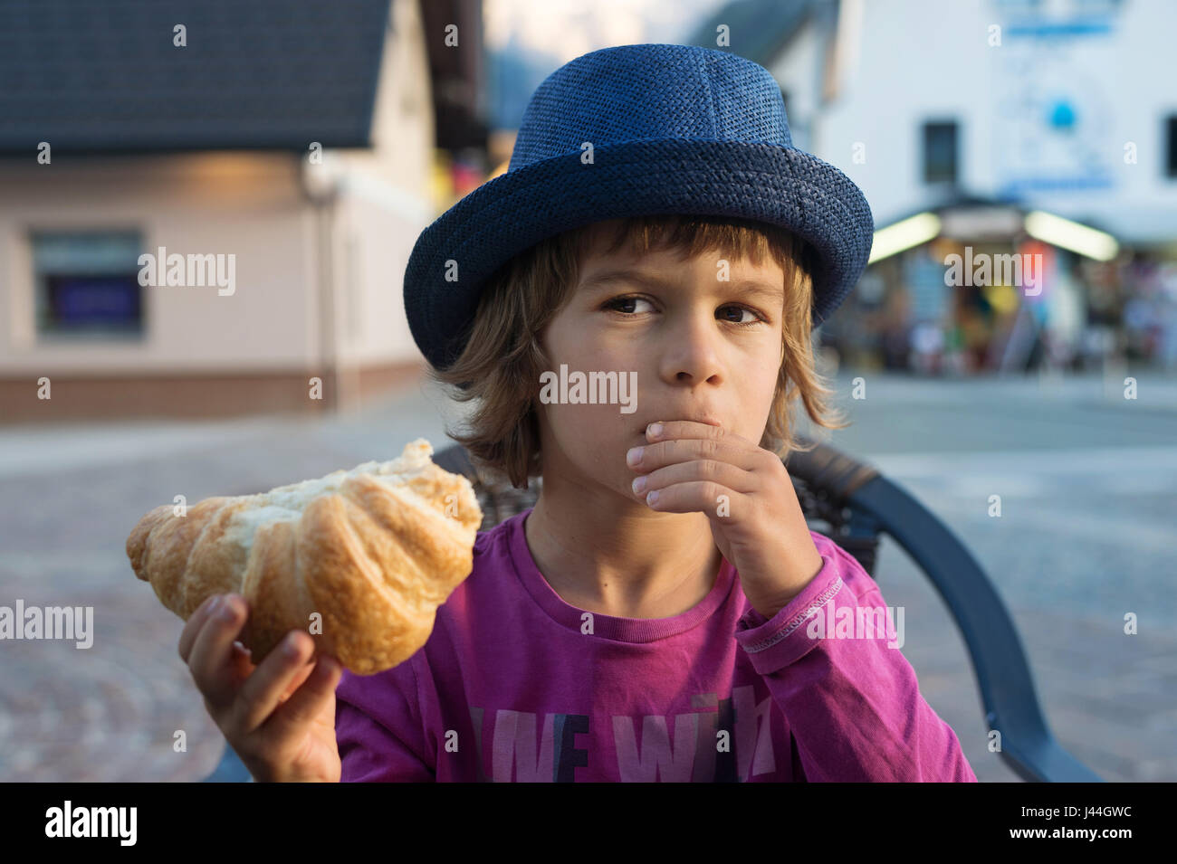 Cute little boy with blue hat sitting at the table eating tasty croissant. Stock Photo