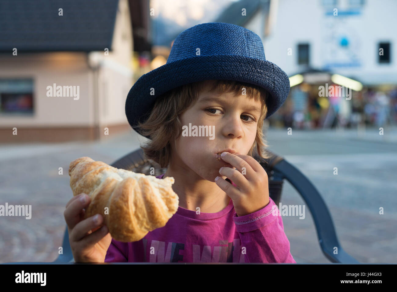 Cute little boy with blue hat sitting at the table stuffing croissant into his mouth. Stock Photo