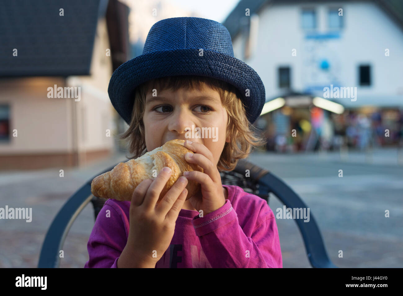 Cute little boy with blue hat sitting at the table eating tasty croissant. Stock Photo