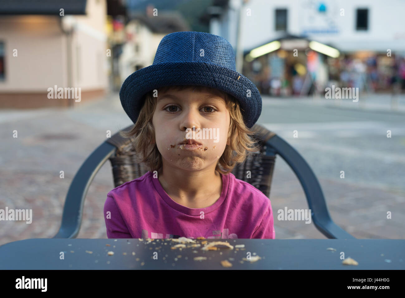 Cute little boy with huge appetite sitting at the table making a mess stuffing croissant into his mouth. Stock Photo