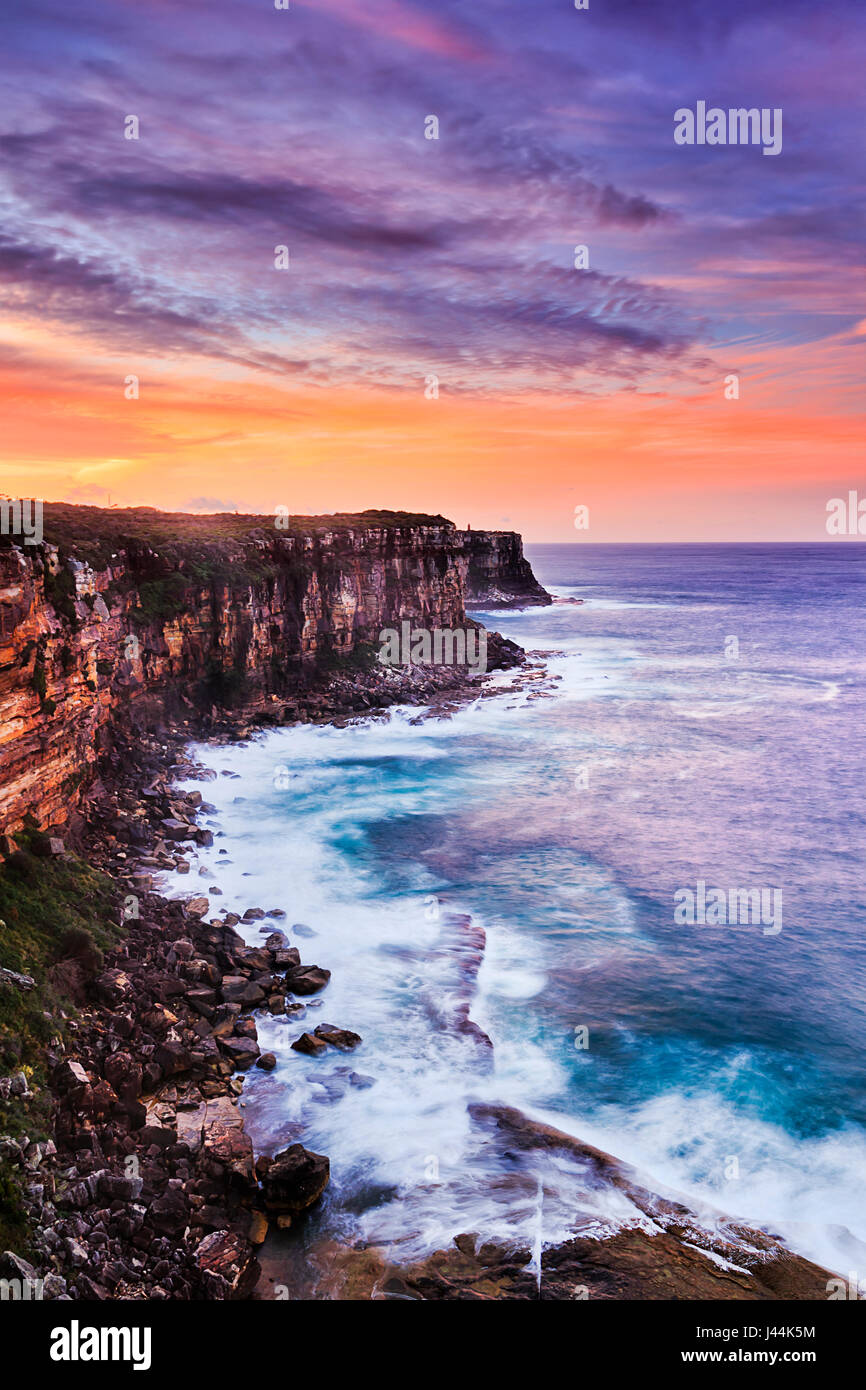 Rugged steep cliffs off Sydney harbour north head facing wild pacific ocean during sunset hours when sky is illuminated in red colour and waves are bl Stock Photo