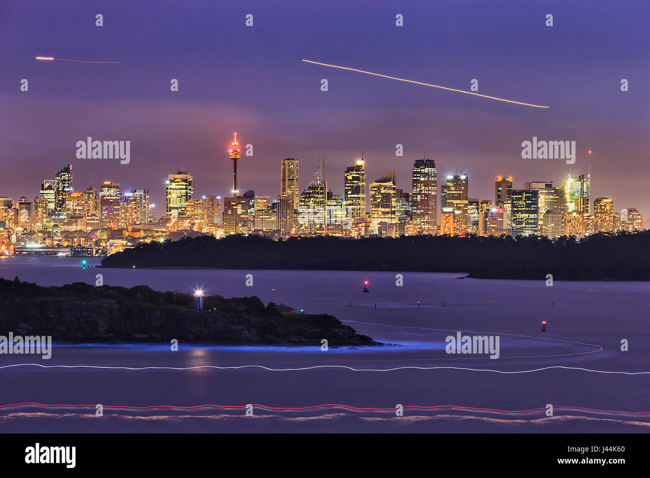 Cityline of Sydney CBD over harbour blurred waters and sandstone shore heads at sunset when the city is brightly illuminated. Stock Photo