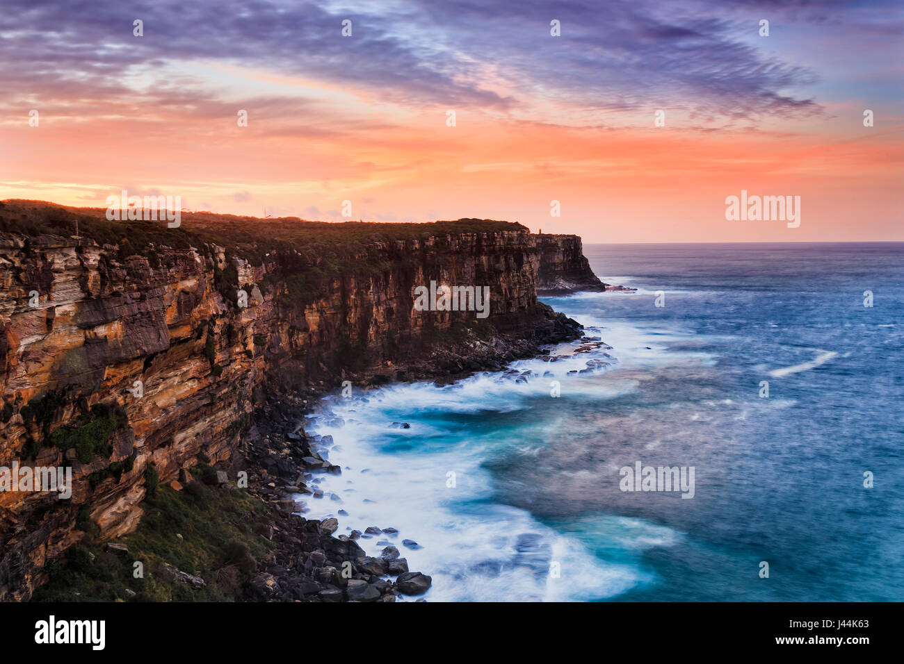 Steep cliffs of rugged sydney coast from North head entrance to Sydney harbour at sunset. Red illuminated sky reflects in blurred waters of surfing pa Stock Photo