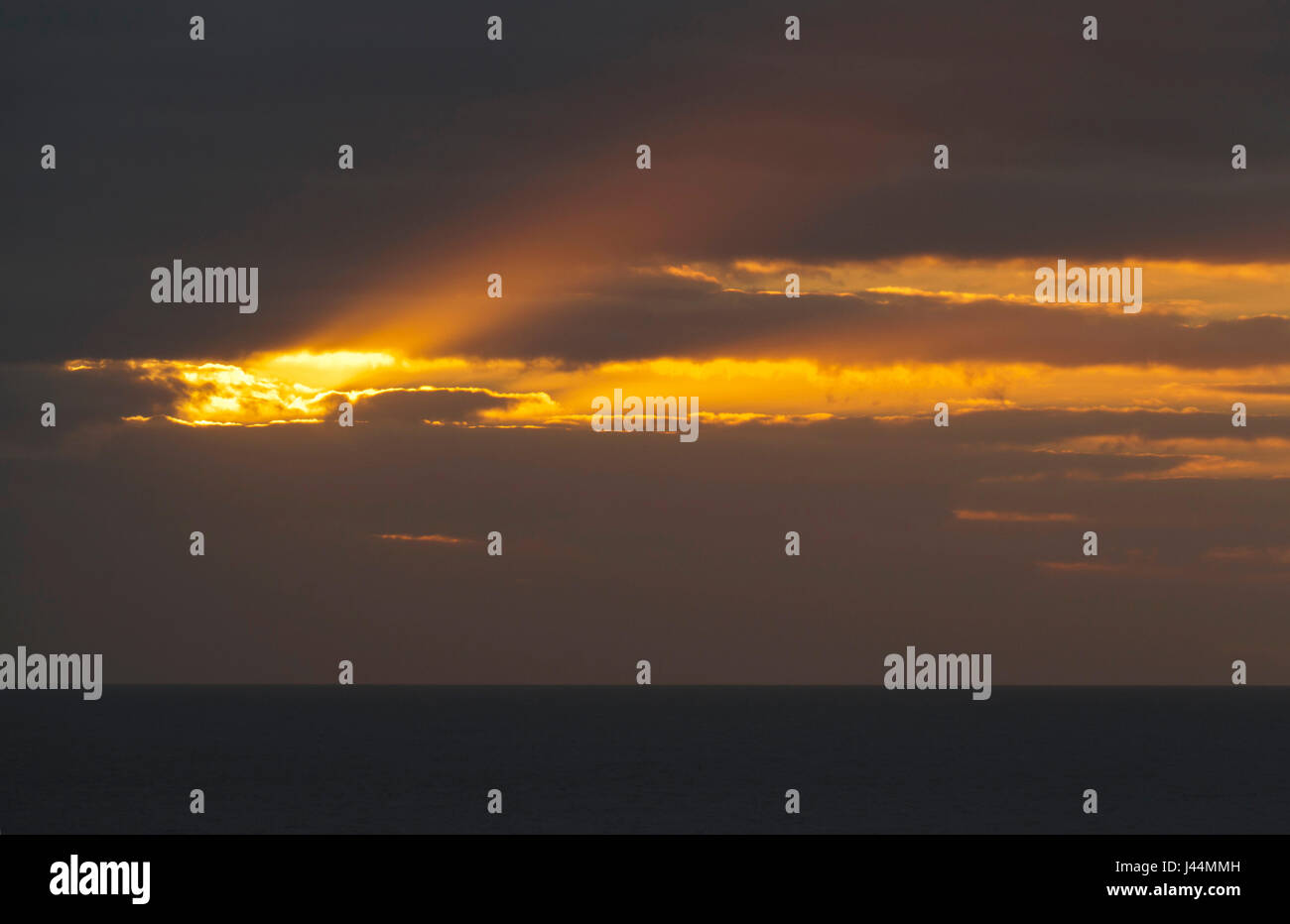 A bright, golden, heavenly shaft of light breaks through an opening in the clouds over a dark sea around sunset Stock Photo