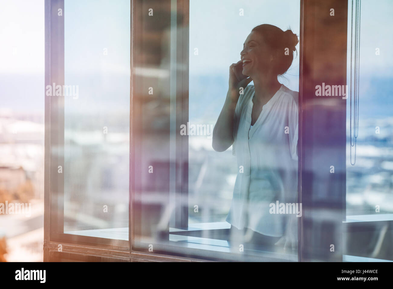 Smiling mature businesswoman standing inside office building and talking on cell phone. Female executive standing by glass window using mobile phone. Stock Photo