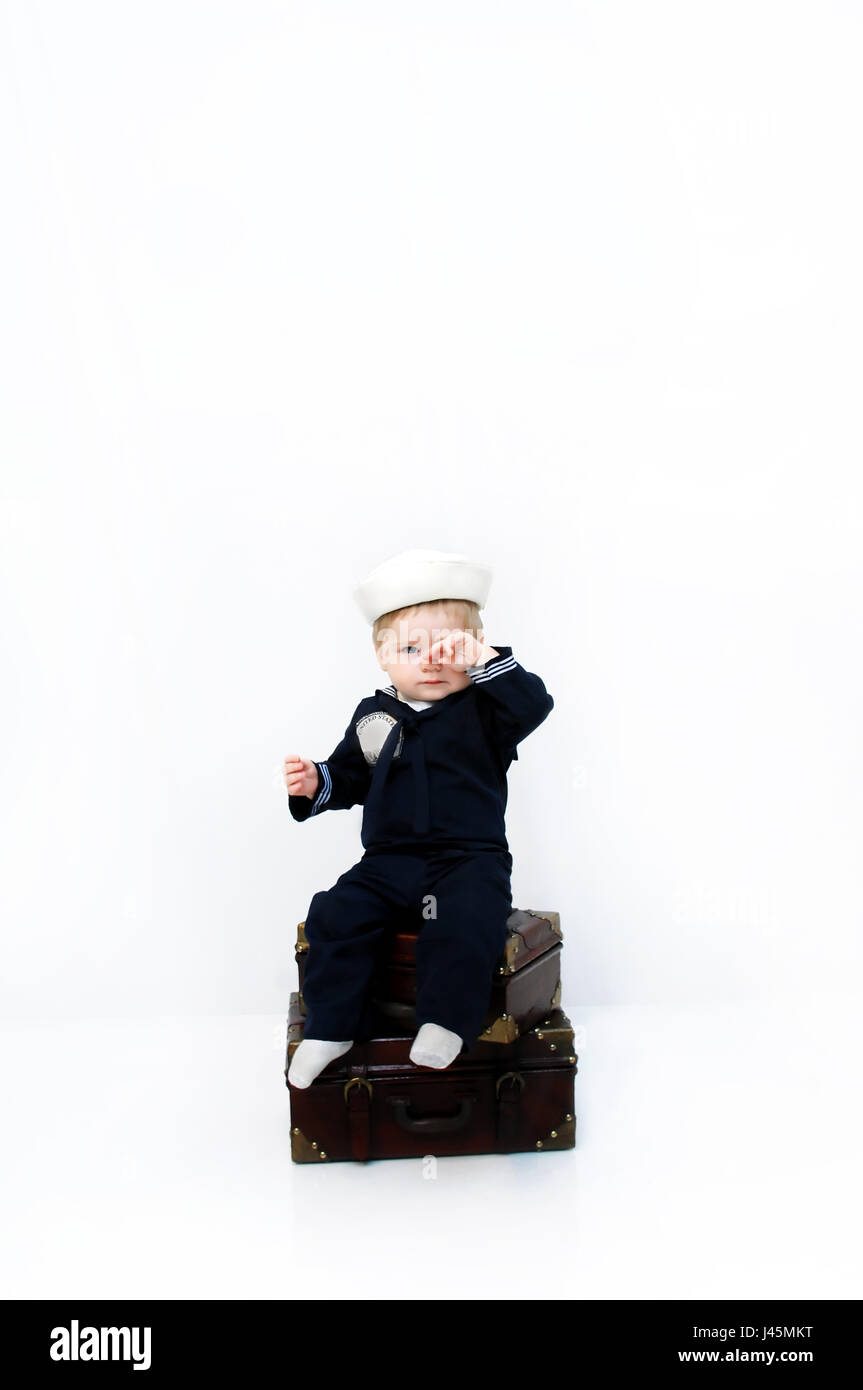 Little baby wearing a naval costume rubs his eyes.  He is sad to be leaving his family.  He is sitting on his luggage waiting to leave. Stock Photo