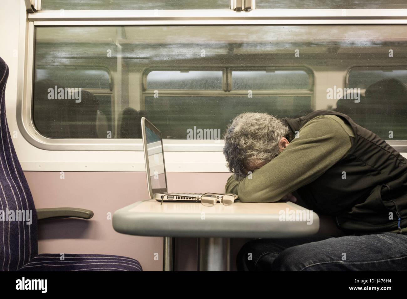 Train UK. Mature man with laptop sleeping at window seat on empty train with rain on window. England. United Kingdom Stock Photo