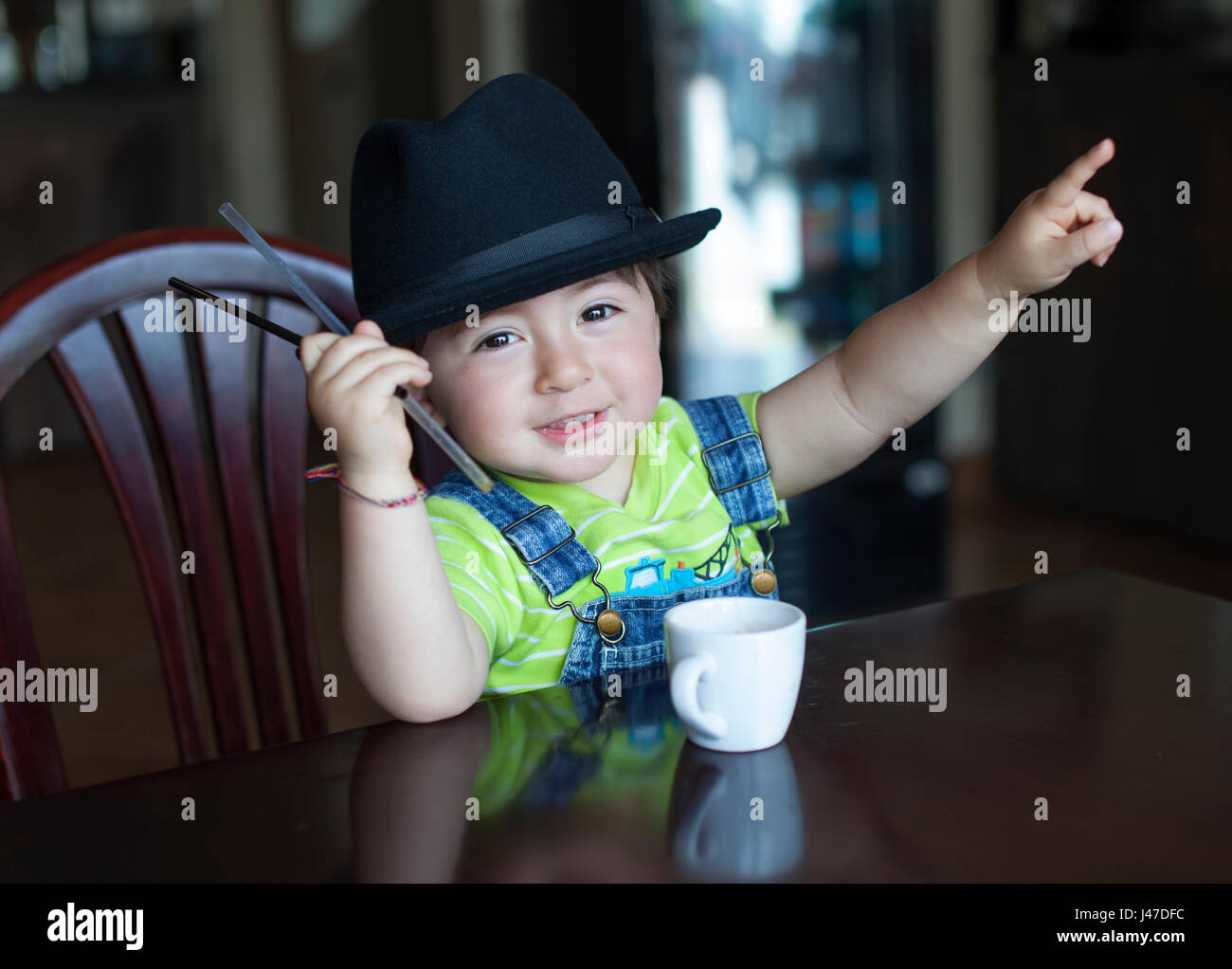 Cute little boy with brown hair wearing a black fedora hat and blue denim jean overalls a yellow shirt with a white porcelain coffee tea cup Stock Photo