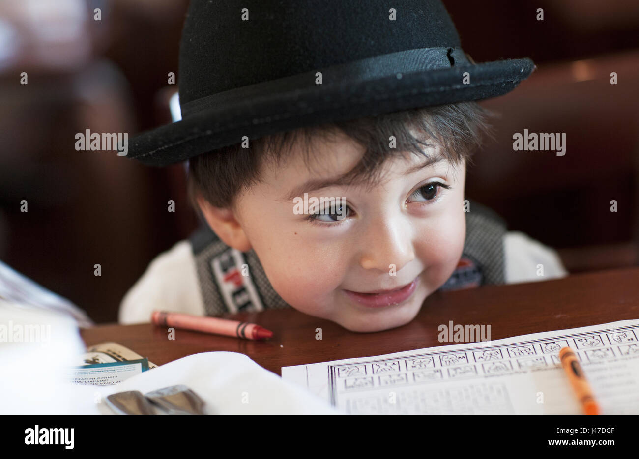 Cute little mixed race Asian Caucasian boy with chubby cheeks wearing a black fedora hat leaning his chin on a table with crayola crayons around him Stock Photo