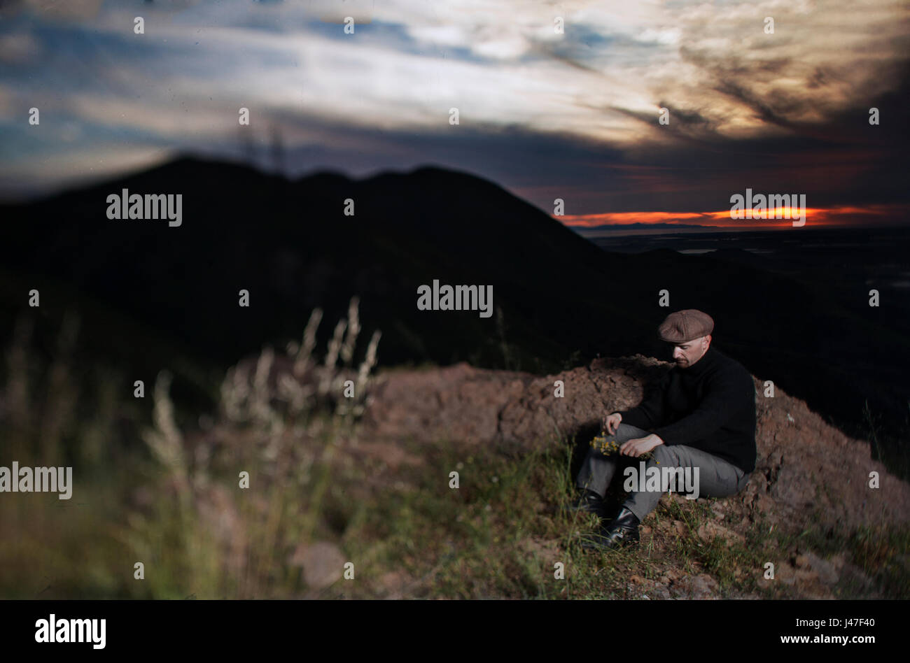 Man with red goatee wearing black turtleneck fisherman's sweater and brown newsboy cap sitting on a cliff with dramatic sunset and clouds Stock Photo