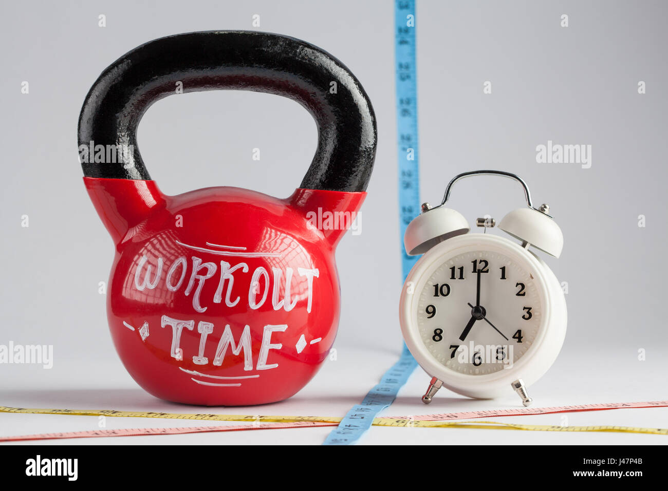 Red kettlebell with Workout Time lettering, traditional alarm clock, and measuring tape isolated on white with copy space. Time to work out, healthy l Stock Photo
