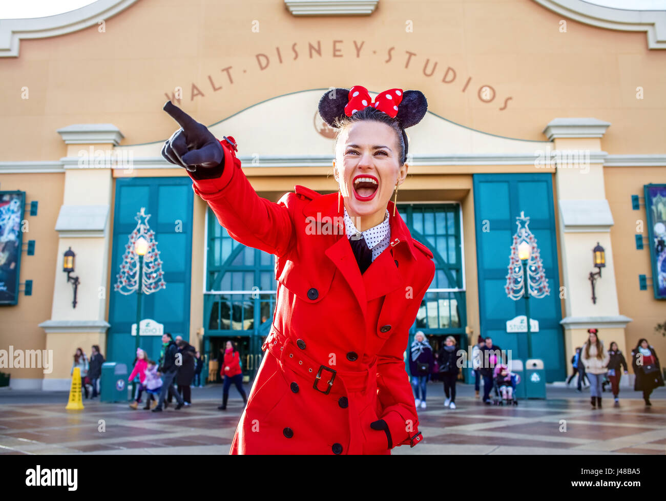 DISNEYLAND, FRANCE - DECEMBER, 8, 2016:. happy modern woman in red trench coat in the front of Disney Studio 1 pointing at something Stock Photo