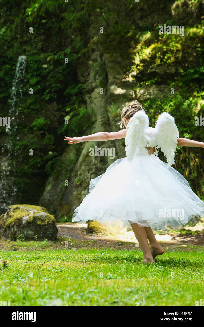 A girl with blond hair wearing an angel costume with feather wings walks on lush grass in a park; Oregon, United States Stock Photo