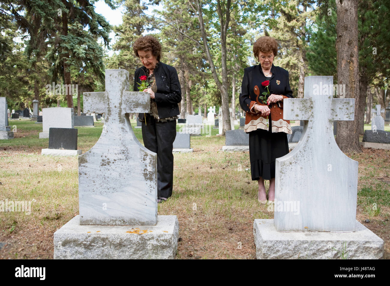 Two Women Holding Single Red Roses Standing At Two Graves Side By Side In A Cemetery; Edmonton, Alberta, Canada Stock Photo