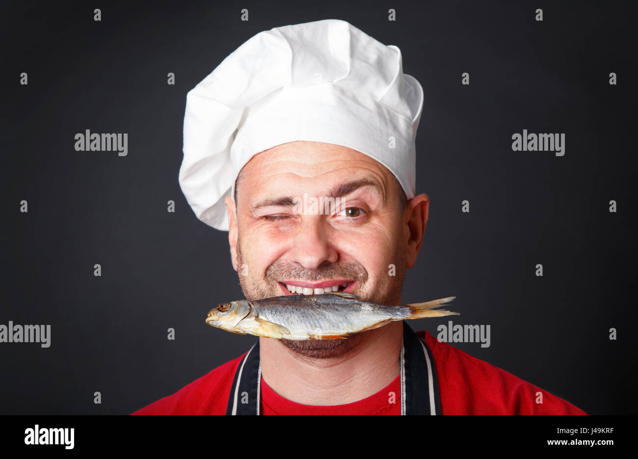 Funny cook in a hat with a dry fish in his teeth in the studio on a black background. Stock Photo
