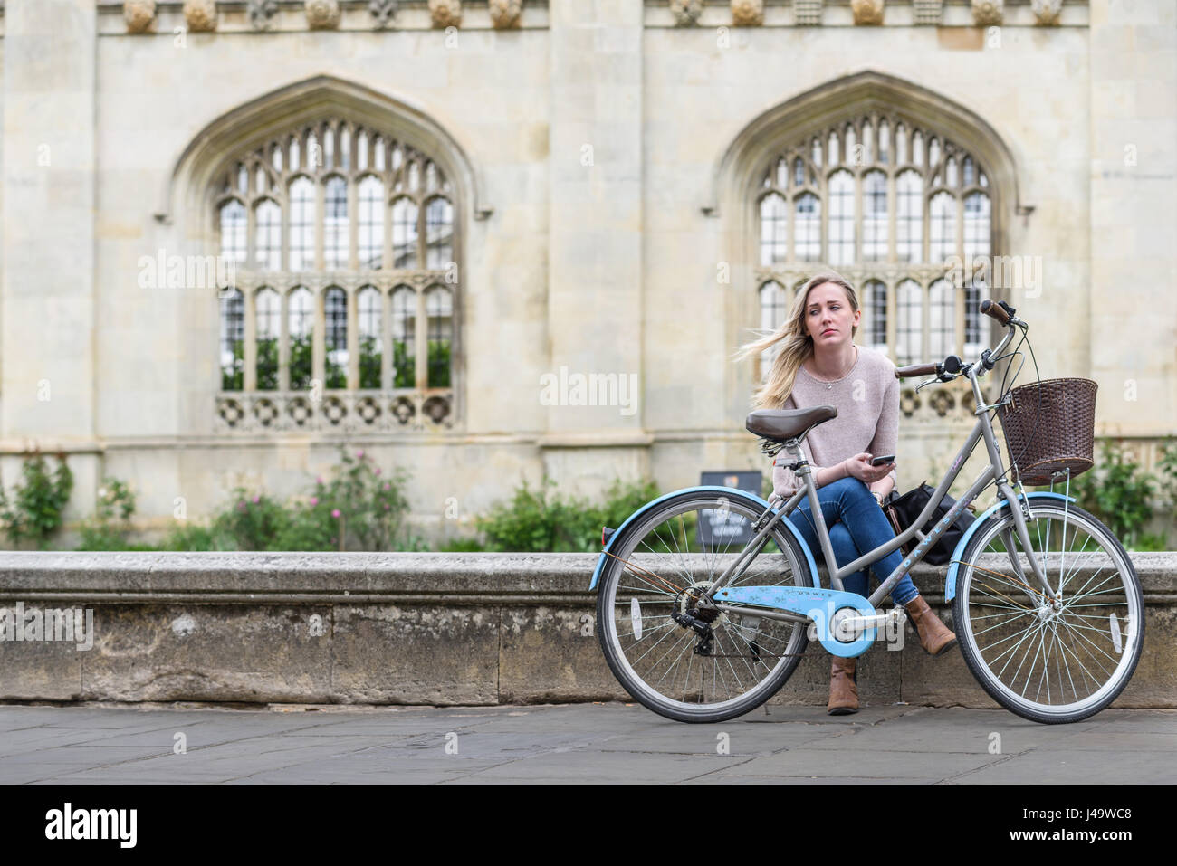Sad student with her bicycle  sits on a low wall in front of King's college at the university of Cambridge, England, Britain. Stock Photo