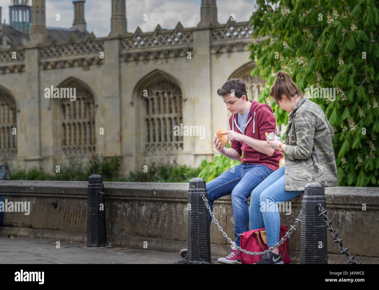 A couple of students sit eating their lunch on a low wall outside the front of King's college at the university of Cambridge, England, Britain. Stock Photo
