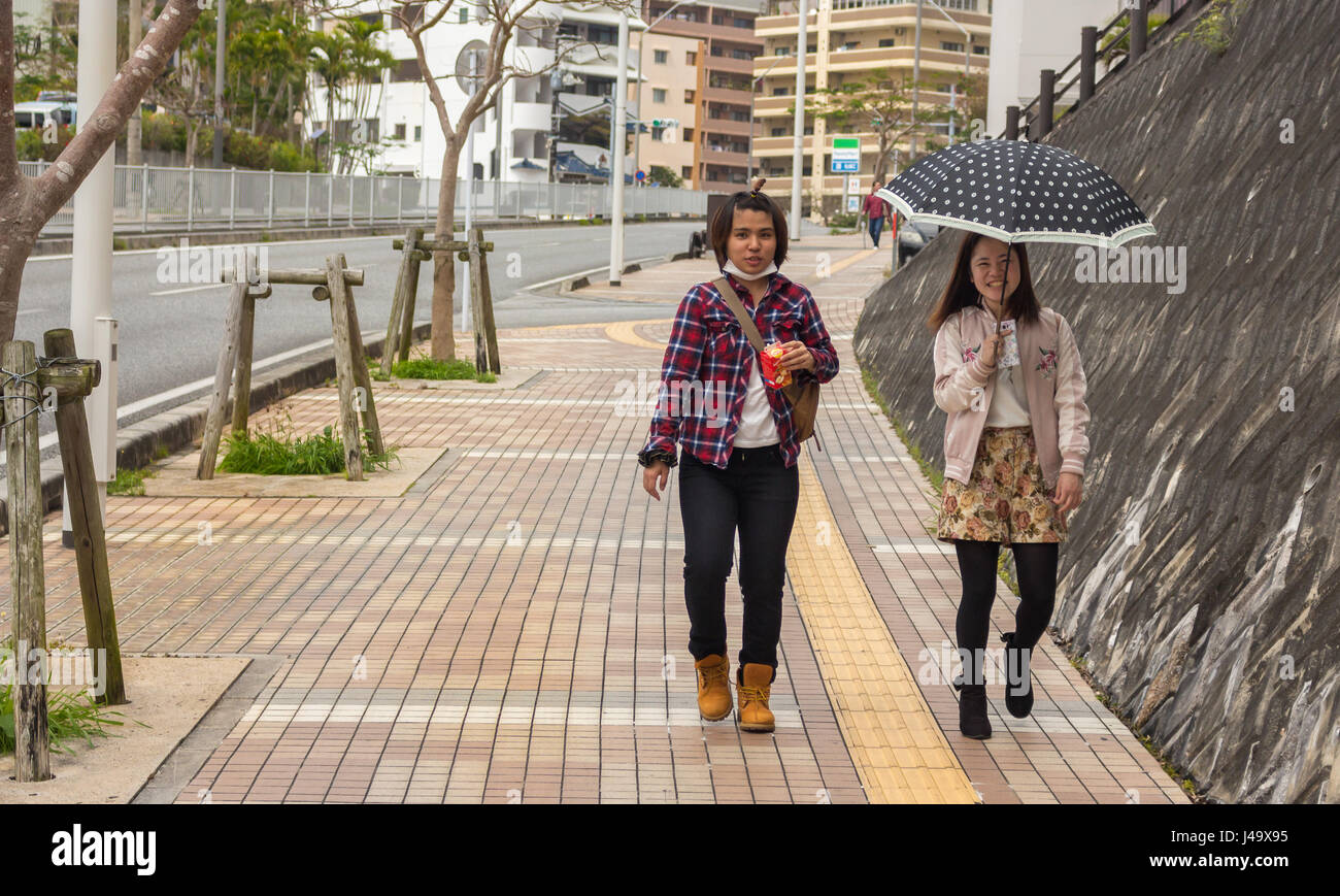 Okinawa, Japan - March 24th, 2017: a japanese girl with a surgical mask and holding an apple juice and another girl holding a mobile and an umbrella w Stock Photo