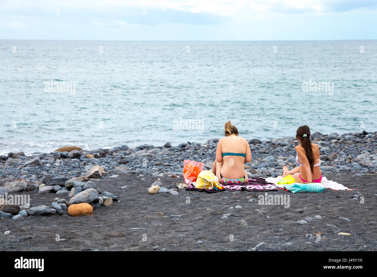 Two women sitting on a black send, volcanic beach, looking out to sea in Tenerife, Spain Stock Photo