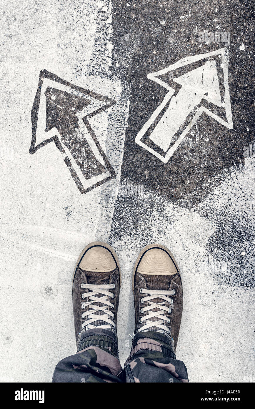 Young man facing decision making situation in life, youth generation Y male wearing worn gray sneakers standing on the street over arrows pointing to  Stock Photo