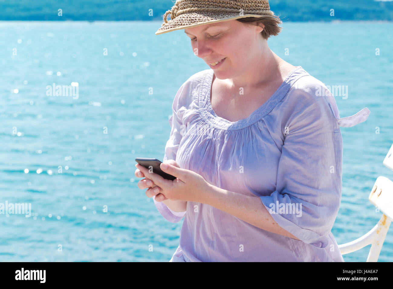 Female tourist with straw hat using mobile phone for communication at seaside on summer holiday vacation Stock Photo