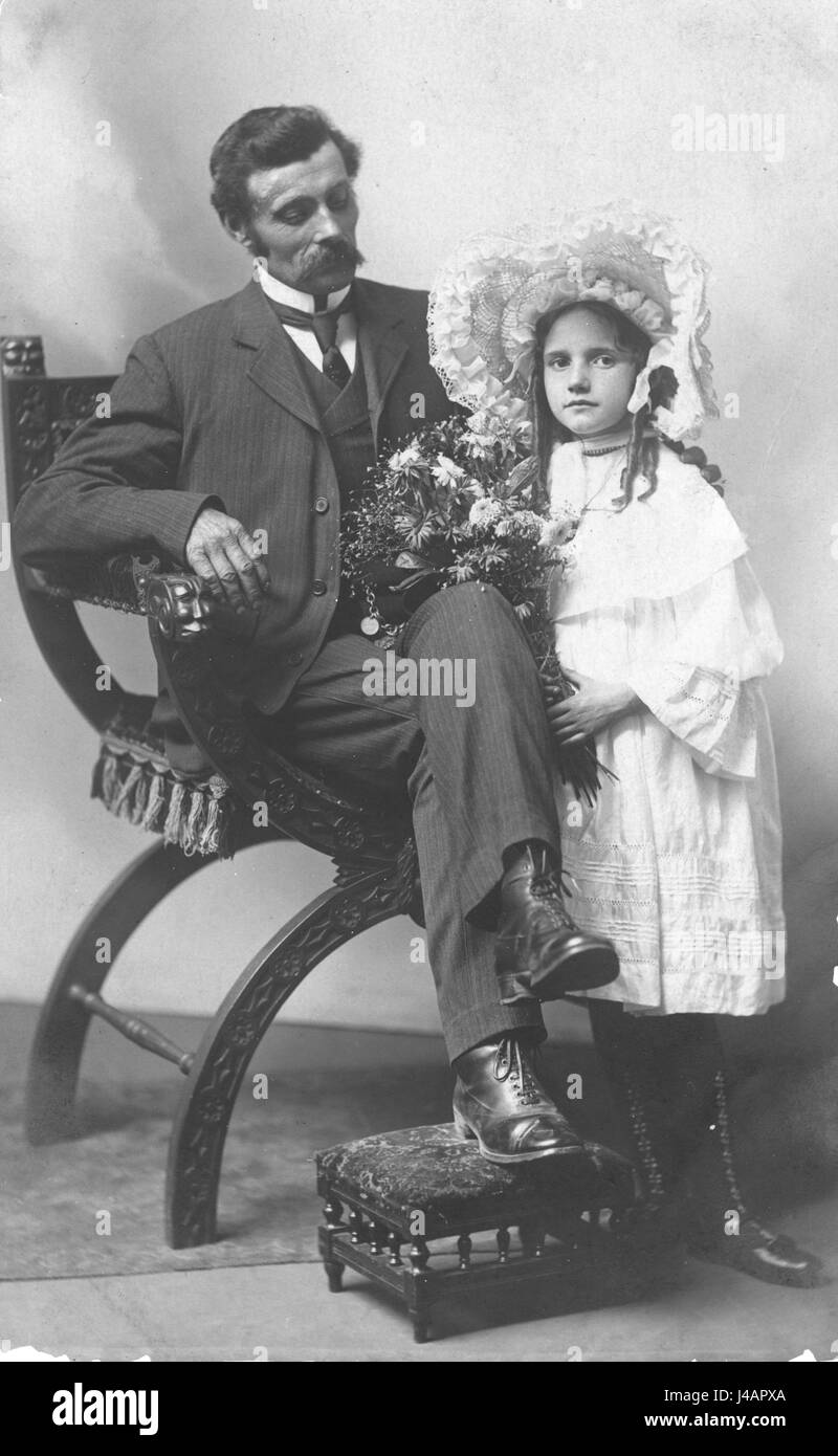 Father proudly gazes at a young girl, presumably his daughter. The girl is finely dressed in a fancy white dress, and she wears a wonderful, big white bonnet.  The man sits on an 1800s era chair, carefully hand carved, which has faces carved on the chair's arm, where the man's hand rests. The chair may be in a Greek revival style. It has a curved seat with tasseled pad for comfort.  Man's foot rests on a great and fancy footstool, hassock.   To see my other child-related images, Search:  Prestor  vintage  kids Stock Photo
