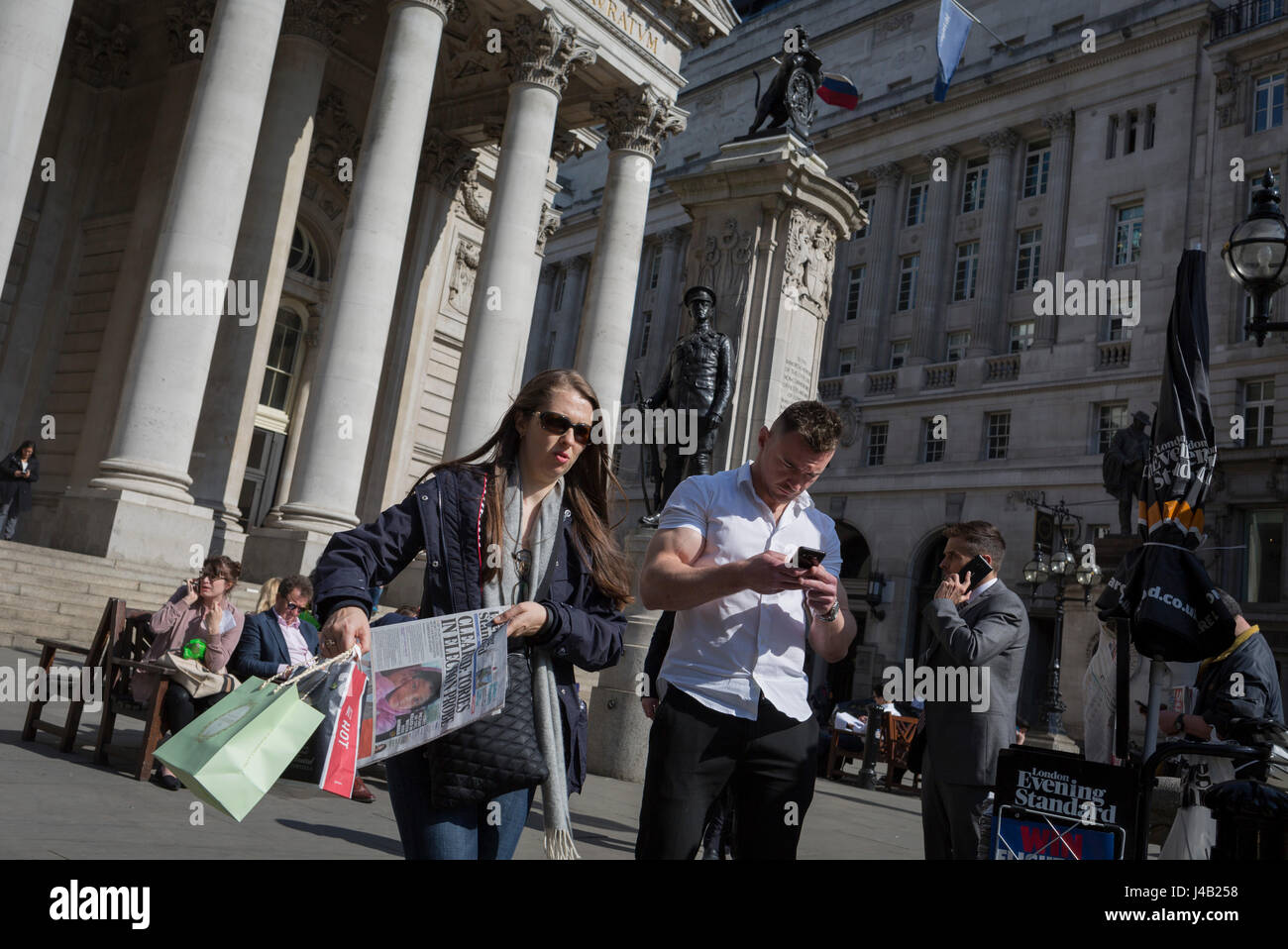 As a woman carries her copy of the Evening Standard newspaper, a muscular young man checks messages below the classical architecture of Royal Exchange and the WW1 war memorial at Bank Triangle, on 10th May 2017, in the City of London, England. Stock Photo