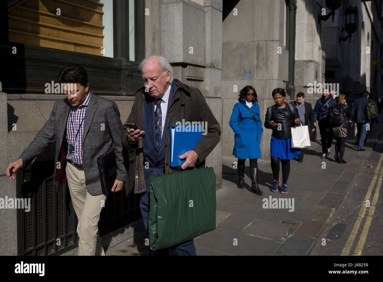 As two women in the background walk in matching blue clothing, an elderly gentleman carries a folder in the same colour, on 10th May 2017, in the City of London, England. Stock Photo