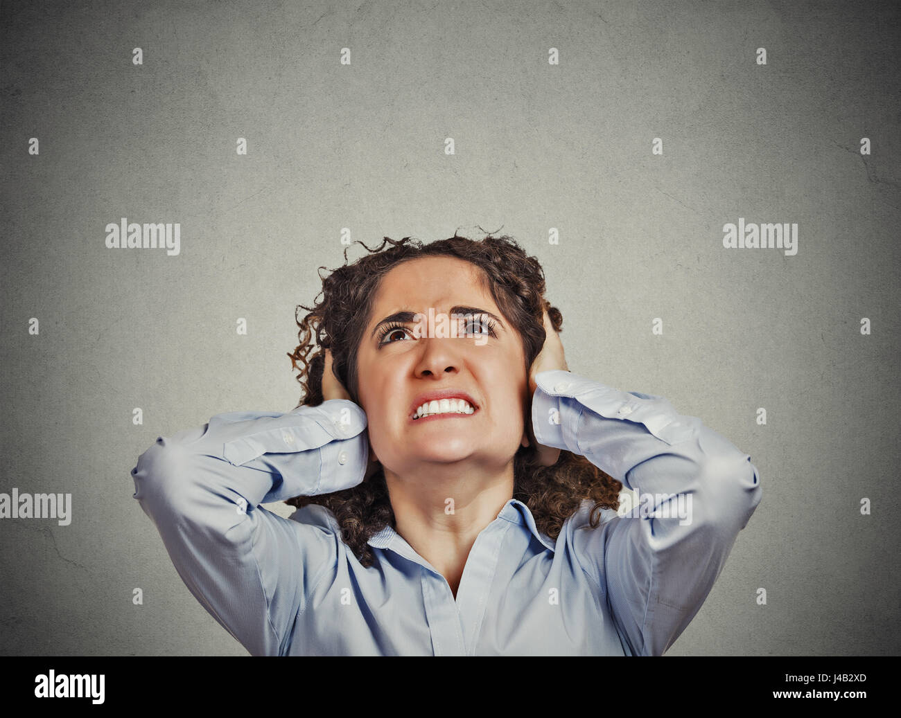 Closeup portrait young angry unhappy stressed woman covering her ears looking up stop making loud noise it's giving me headache isolated grey wall bac Stock Photo