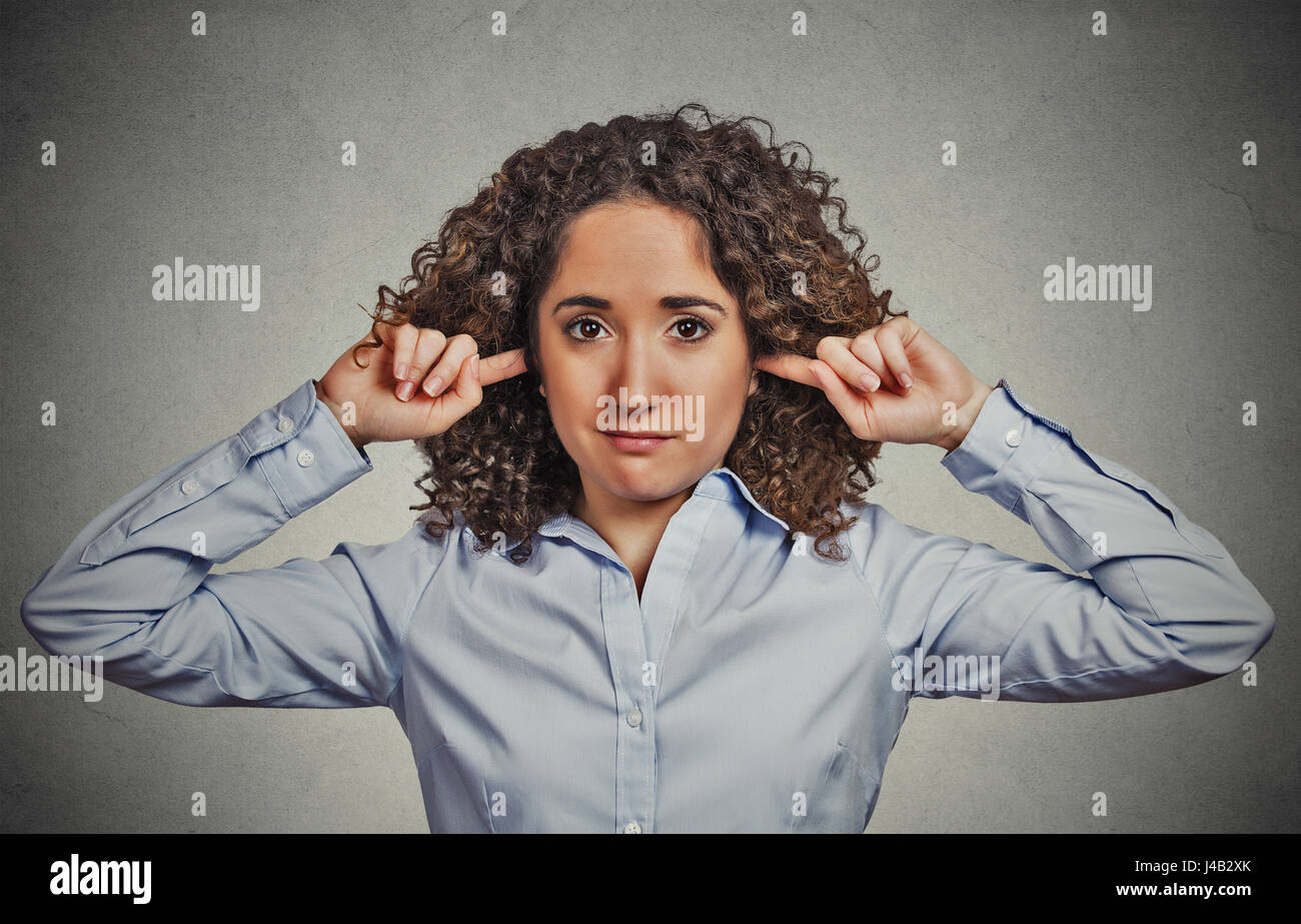 Closeup portrait young angry, unhappy woman covering closed ears looking at you annoyed by loud noise giving her headache ignoring isolated grey wall  Stock Photo
