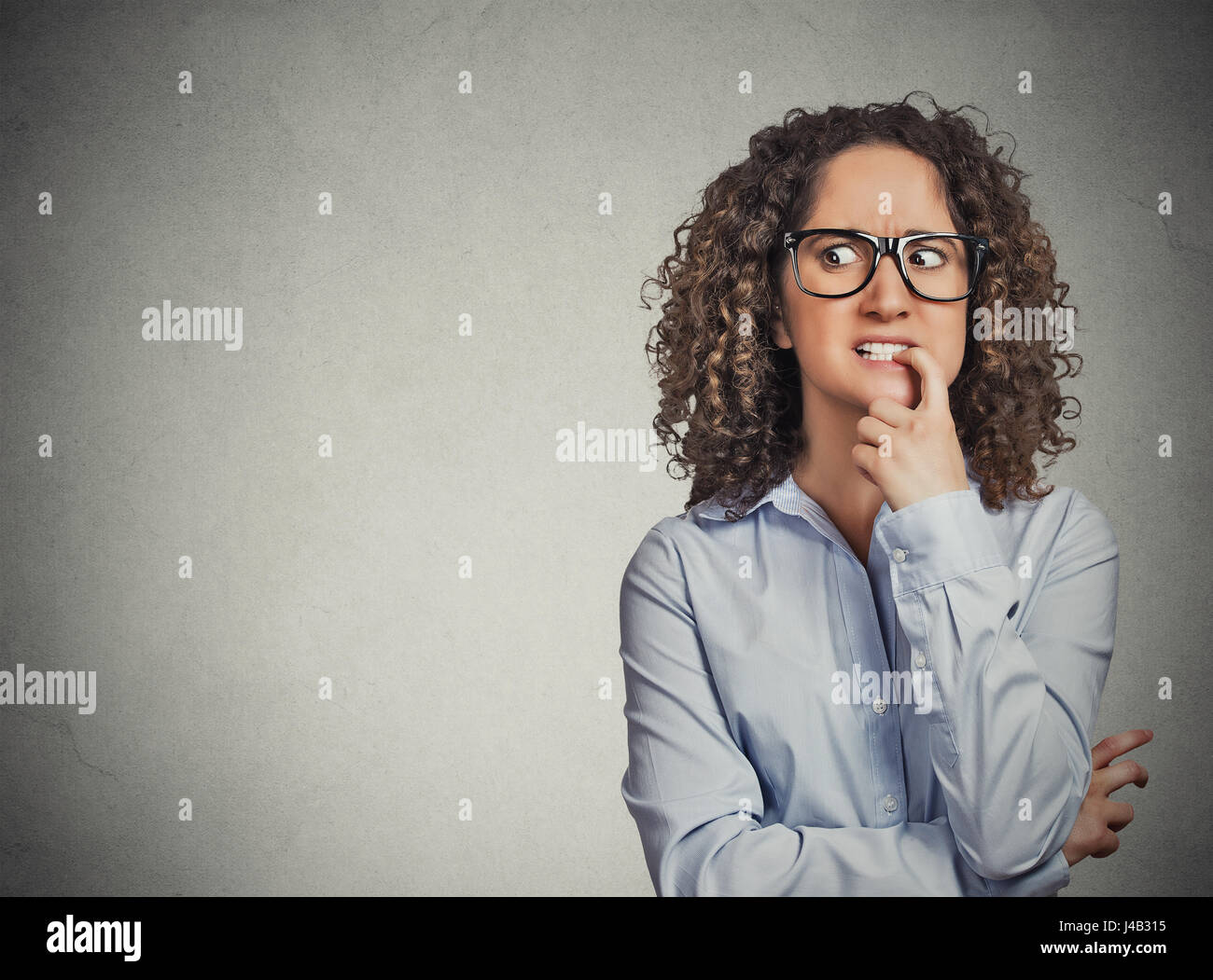 Closeup portrait nervous woman with glasses biting her fingernails craving for something, anxious isolated grey wall background with copy space. Negat Stock Photo