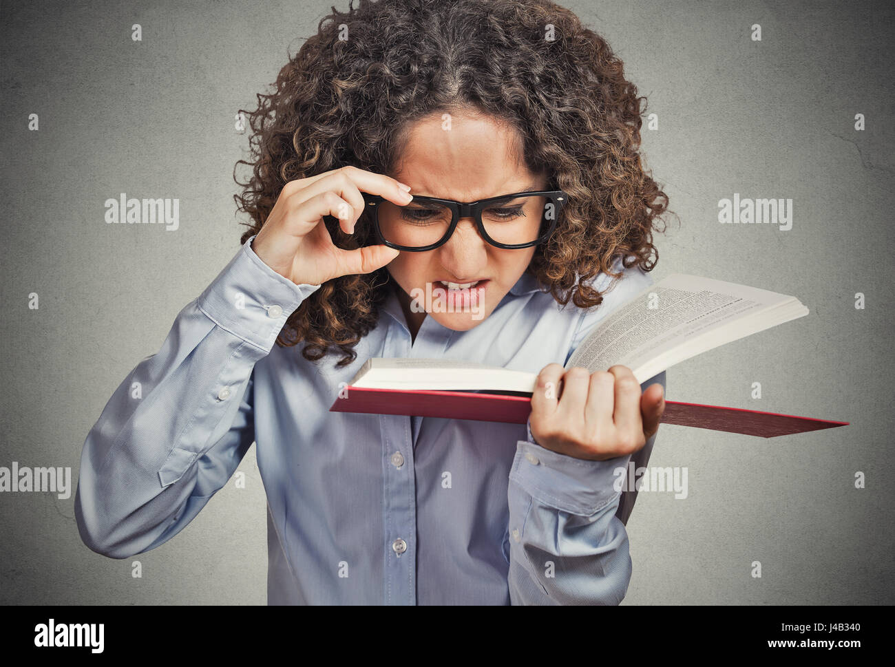 Closeup portrait young woman with eye glasses trying to read book, having difficulties seeing text, bad vision sight problem isolated grey background. Stock Photo