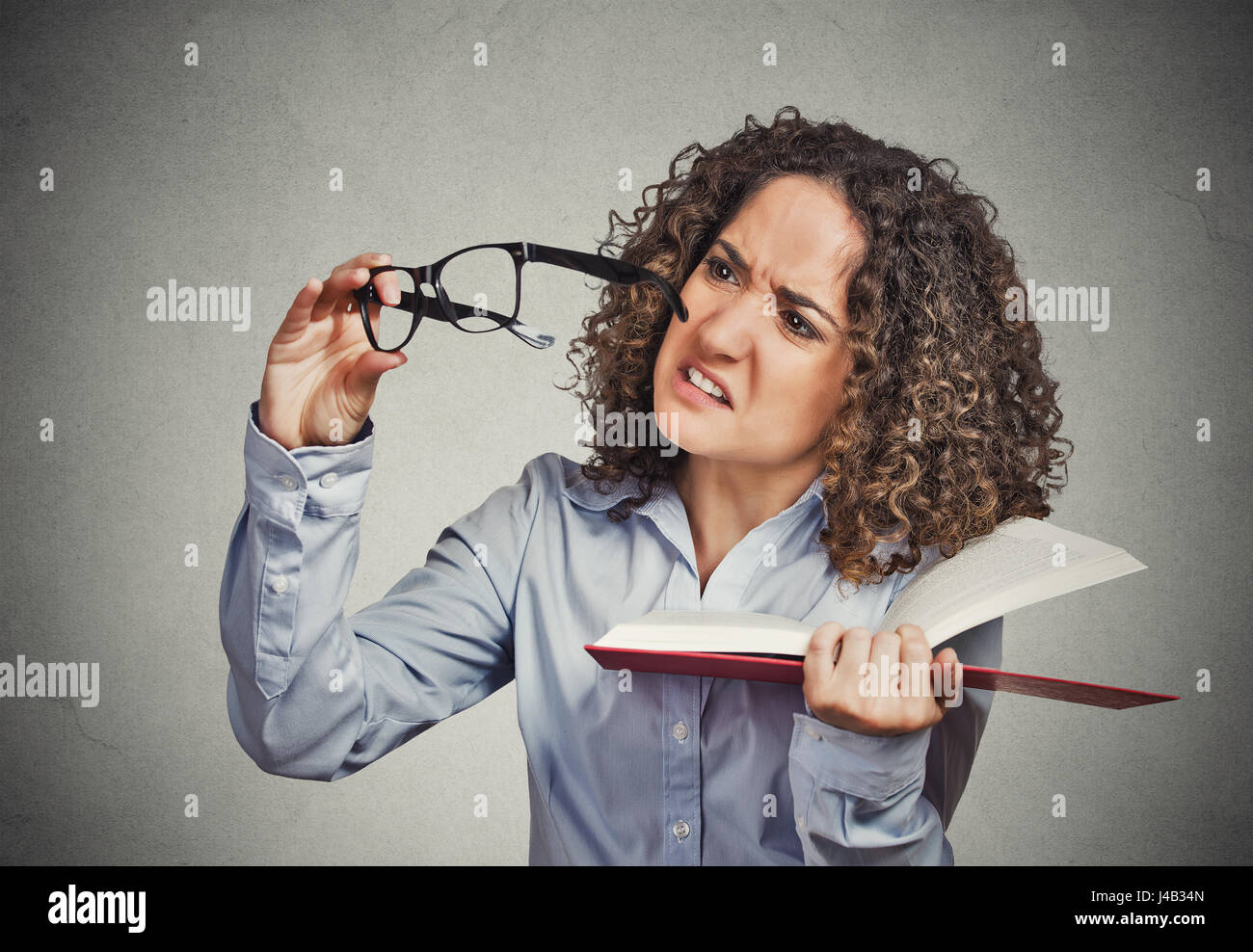 Closeup portrait young lady, woman can't see read book has vision problems wrong glasses prescribed upset isolated grey wall background. Human emotion Stock Photo