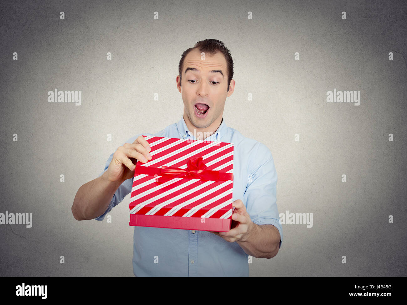 Closeup portrait happy super excited surprised young man about to open unwrap red gift box isolated grey wall background enjoying his present. Positiv Stock Photo