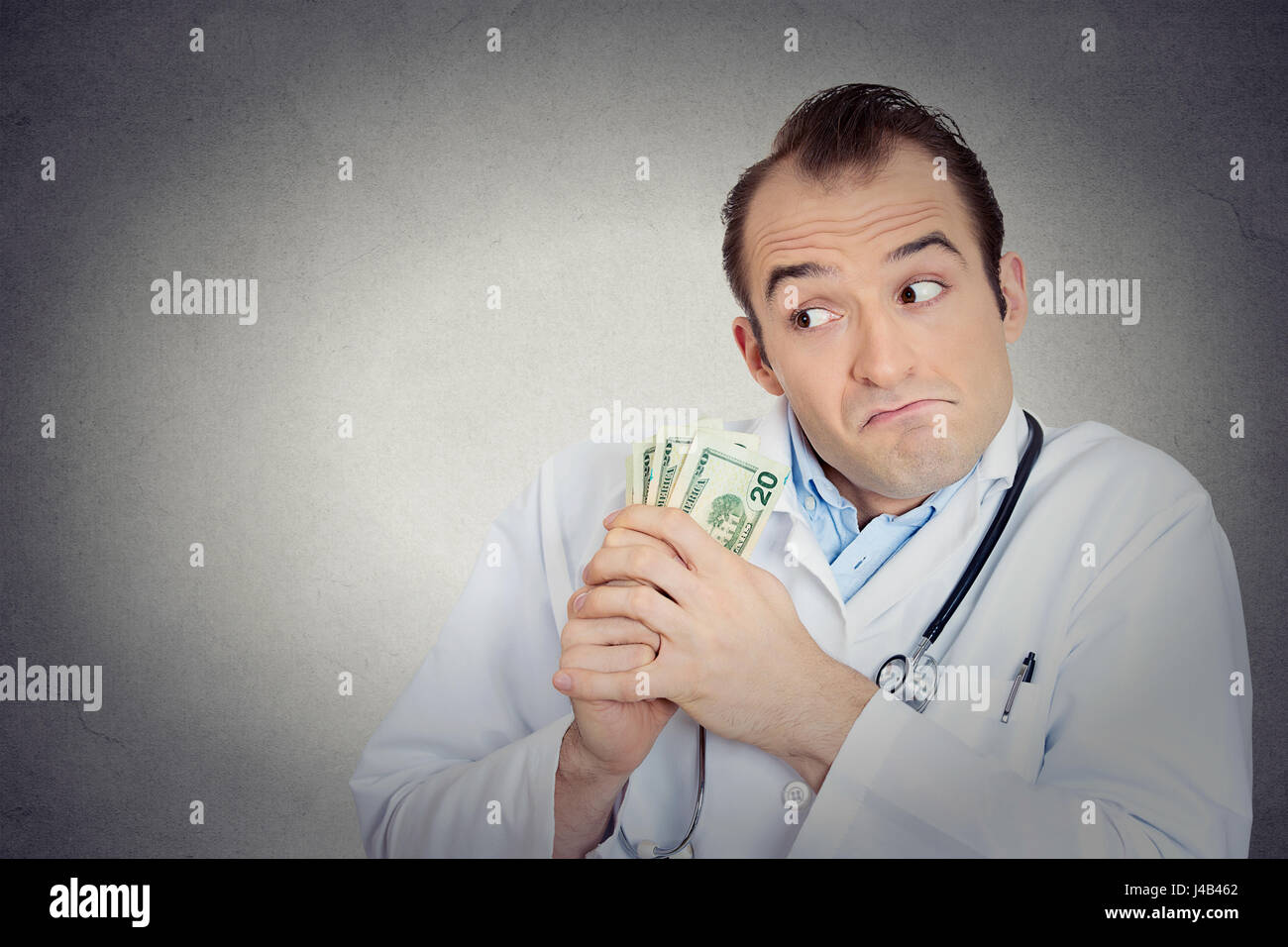 Closeup portrait grumpy greedy miserly health care professional, male doctor holding, protecting his money dollars in hand isolated grey wall backgrou Stock Photo