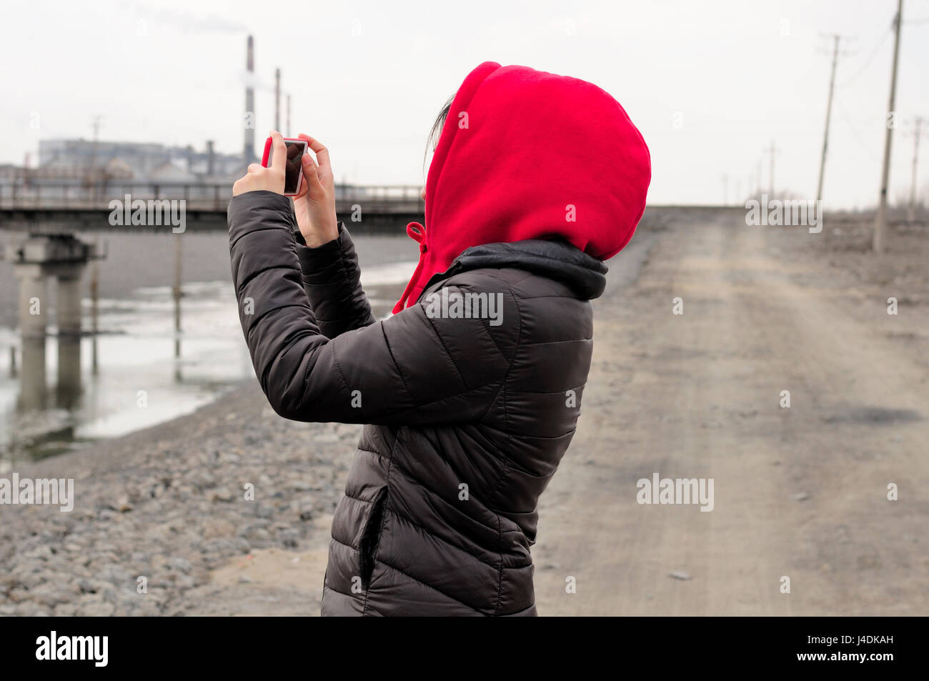A chinese woman wearing a red hoodie taking pictures with a smart phone near a chinese ethanol production factory along a dirt road in the city of Zha Stock Photo