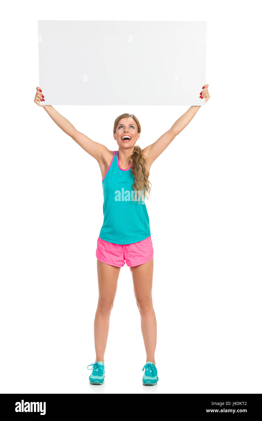 Excited woman in sports clothes holding white empty placard over her head. Full length studio shot isolated on white. Stock Photo