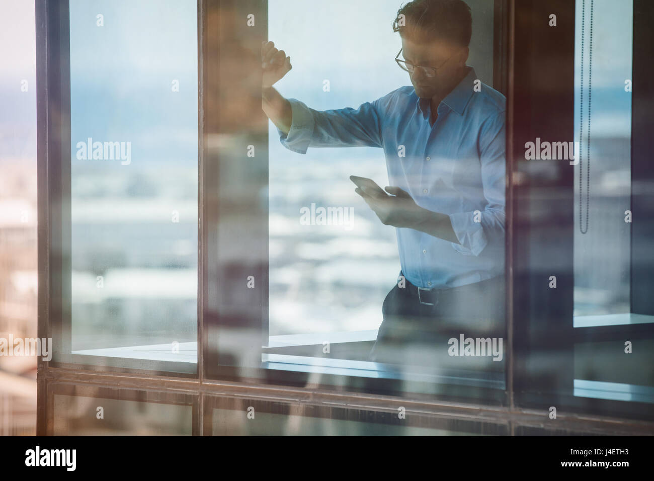 Mature businessman standing by window and reading text message on his mobile phone. Entrepreneur standing inside office building and using smart phone Stock Photo