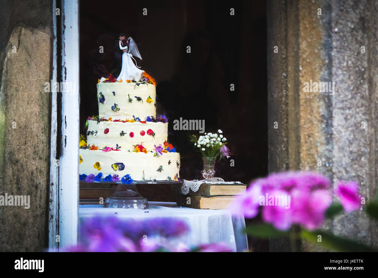 Pretty wedding cake with white icing and flowers topped with figurines of the bride and groom. Pink and purple flowers in the foreground, blurred back Stock Photo