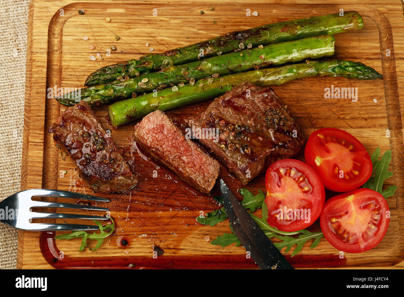 Cut slices of grilled juicy medium cooked beefsteak served on wooden board with fresh green rocket salad, asparagus and red cherry tomatoes, fork and  Stock Photo